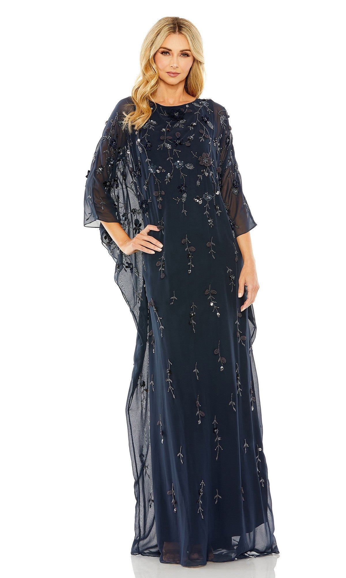 A woman poses gracefully with one hand on her hip, wearing the Mac Duggal 9242 Trapeze Embellished Formal Gown—a floor-length navy evening dress featuring sheer fabric, floral embroidery, and loose sleeves.